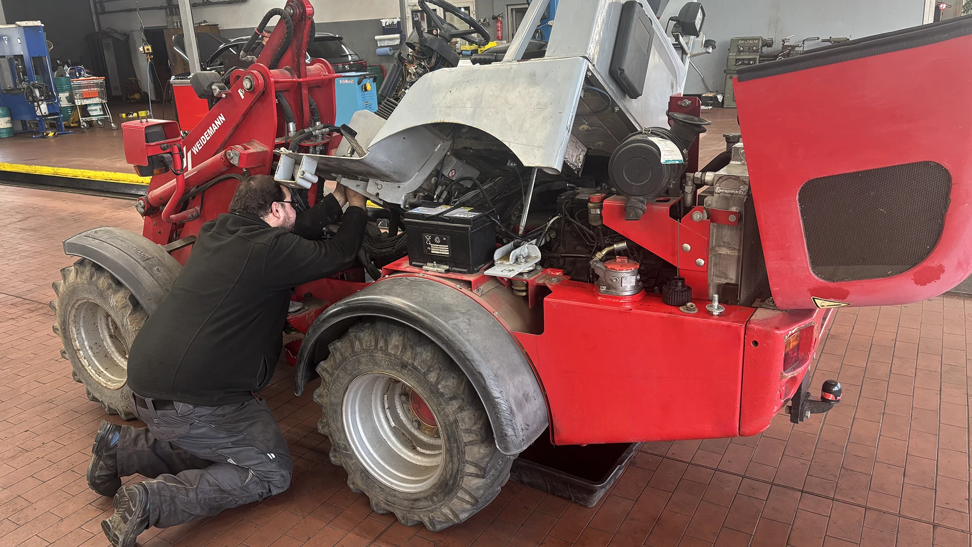 Foto zeigt Reparatur eines Weidemann-Traktors in Werkstatthalle mit davor knieendem Monteur